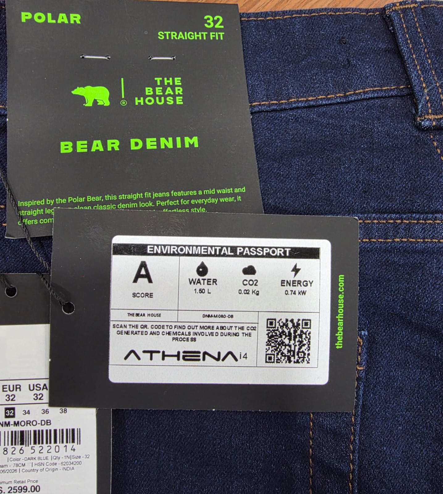 An Athena i4 Environmental Passport hangtag on a pair of The Bear House denim jeans, showing score A, 1.50 litres of water, 0.02 kilograms of CO2 and 0.74 kilowatts of energy